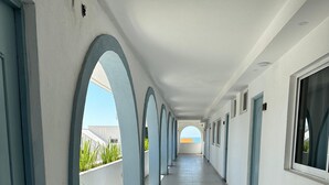 Un largo pasillo con paredes blancas y marcos de puerta azules, que conduce a un balcón con vistas al mar.