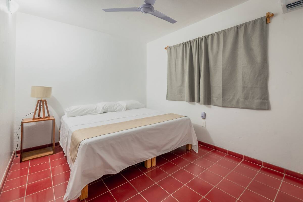 A bedroom with a bed, a nightstand, a lamp, a ceiling fan, and a window with curtains.