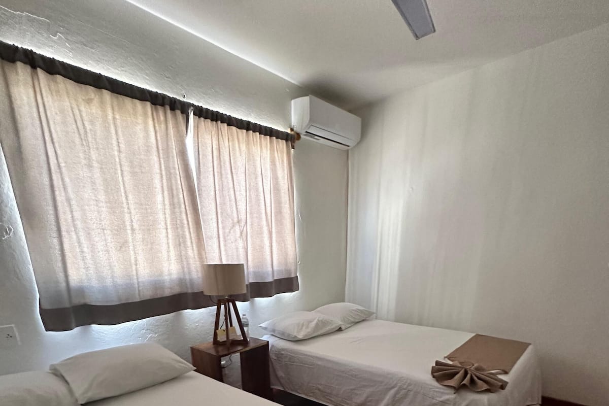 A hotel room with two beds, a ceiling fan, an air conditioning unit, and a window with curtains.