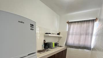 A compact kitchen with a white refrigerator, a microwave, and a sink. The floor is tiled with red and white patterns, and there's a blue trash bin.