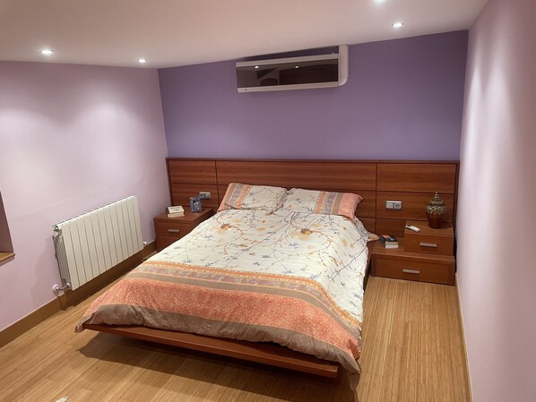 Une chambre à coucher avec un lit en bois, une table de chevet avec une lampe, une petite table à côté du lit avec un livre, et un climatiseur fixé au mur.