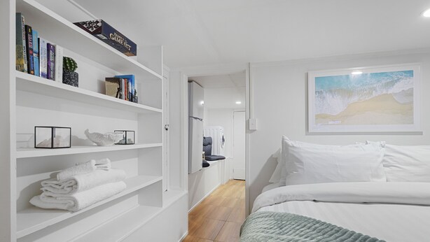 Une chambre à coucher avec un lit, des murs blancs, une bibliothèque remplie de livres et d’objets décoratifs, et un tableau encadré accroché au mur.
