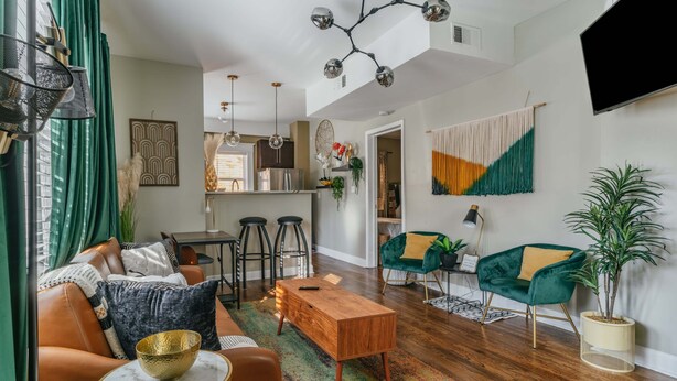 Una sala de estar moderna con un sofá, una mesa de centro y sillas, un área de cocina al fondo y un televisor montado en la pared.