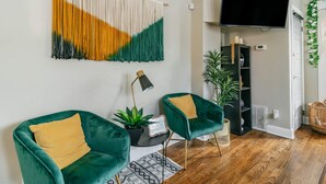 Una sala de estar con dos sillones de terciopelo verde, un tapiz abstracto de gran tamaño, un televisor de pantalla plana y una planta en maceta.