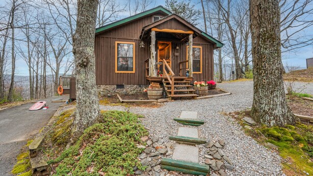 Une cabane rustique aux murs en bois, dotée d’un porche avec des marches et d’une allée de gravier bordée d’arbres.