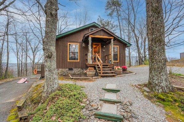 Une cabane rustique aux murs en bois, dotée d’un porche avec des marches et d’une allée de gravier bordée d’arbres.