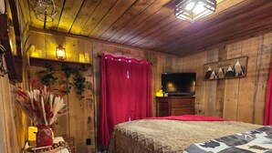 A cozy room with wooden walls, a bed with a patterned blanket, a television on a wooden dresser, and a red curtain. - AUTHENTIC, Rustic, Cute, Cozy and close to everything! Real Cabin in the woods! (Pigeon Forge)