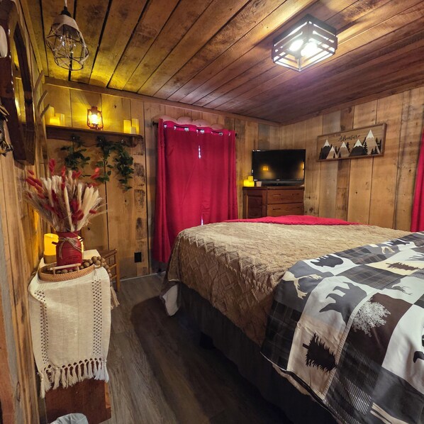 Une pièce chaleureuse aux murs en bois, un lit recouvert d’une couverture à motifs, un téléviseur posé sur une commode en bois et un rideau rouge.