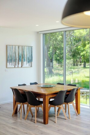 A modern dining area with a large wooden table and black chairs, a large window with a view of greenery, and a piece of abstract art on the wall. - Serenity HG4: 4Br/3.5Ba with Absolute NP Frontage (Halls Gap)