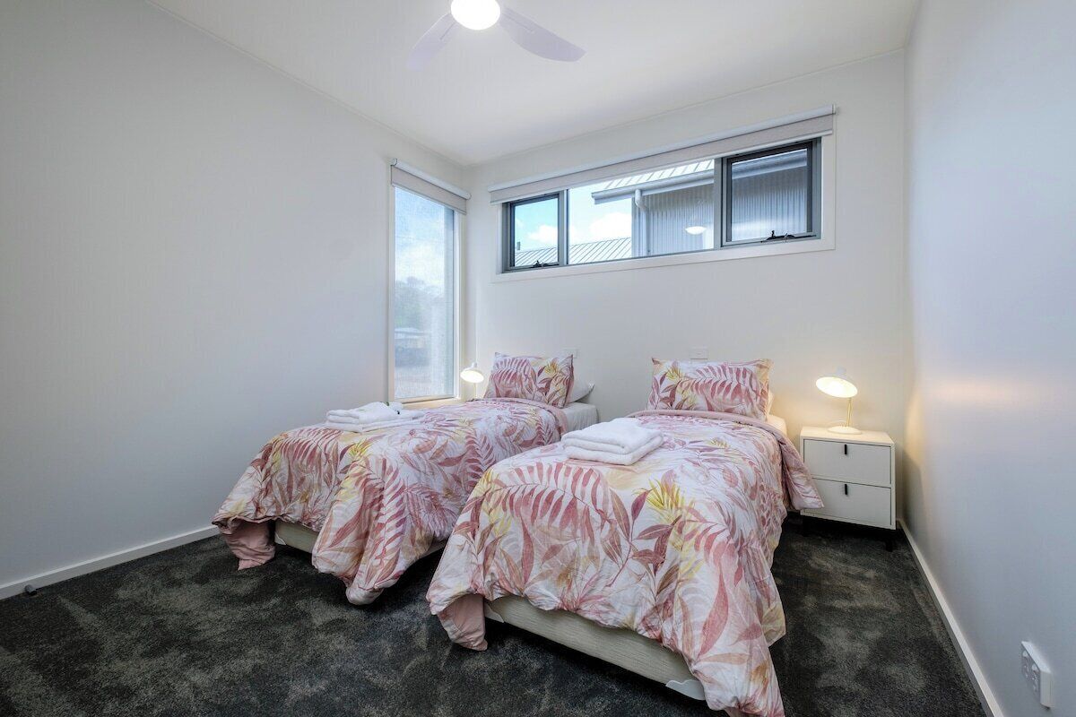 Un dormitorio con dos camas, un ventilador de techo, una ventana con persianas y una mesita de noche con lámpara.