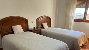 A hotel room with two beds, wooden headboards, a nightstand with a coffee maker, and a window with blinds.