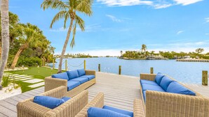 A wooden deck with wicker furniture and blue cushions overlooking a body of water with palm trees and a distant island. - 4 Bed / 4 Bath with Stunning Views of Bimini Bay ! (Anna Maria)