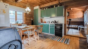 Interior rústico de una cabaña con una mesa de comedor de madera, sillas, una lámpara colgante, una cocina con armarios verdes y un ventanal grande.