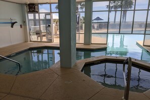 An indoor swimming pool with clear views of the outside, including a beach and palm trees.