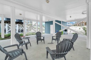 Un área de estar al aire libre cubierta con sillas Adirondack y piso de concreto, con vista a una playa y edificios al fondo.