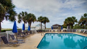 A swimming pool with lounge chairs and umbrellas, surrounded by palm trees and a white fence. - Dandy Sandy: Sleeps 24 - Pets - Pools - Hot Tubs (Gulf Shores)