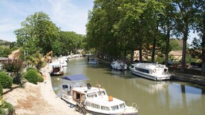 Un canal bordeado de barcos y árboles, con una embarcación atracada que presenta una zona cubierta y una pequeña construcción.