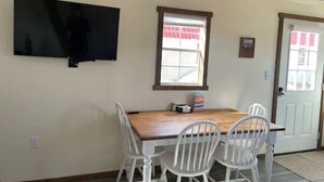 Un coin repas avec une table en bois et des chaises blanches, un téléviseur à écran plat fixé au mur et une fenêtre donnant sur un immeuble.