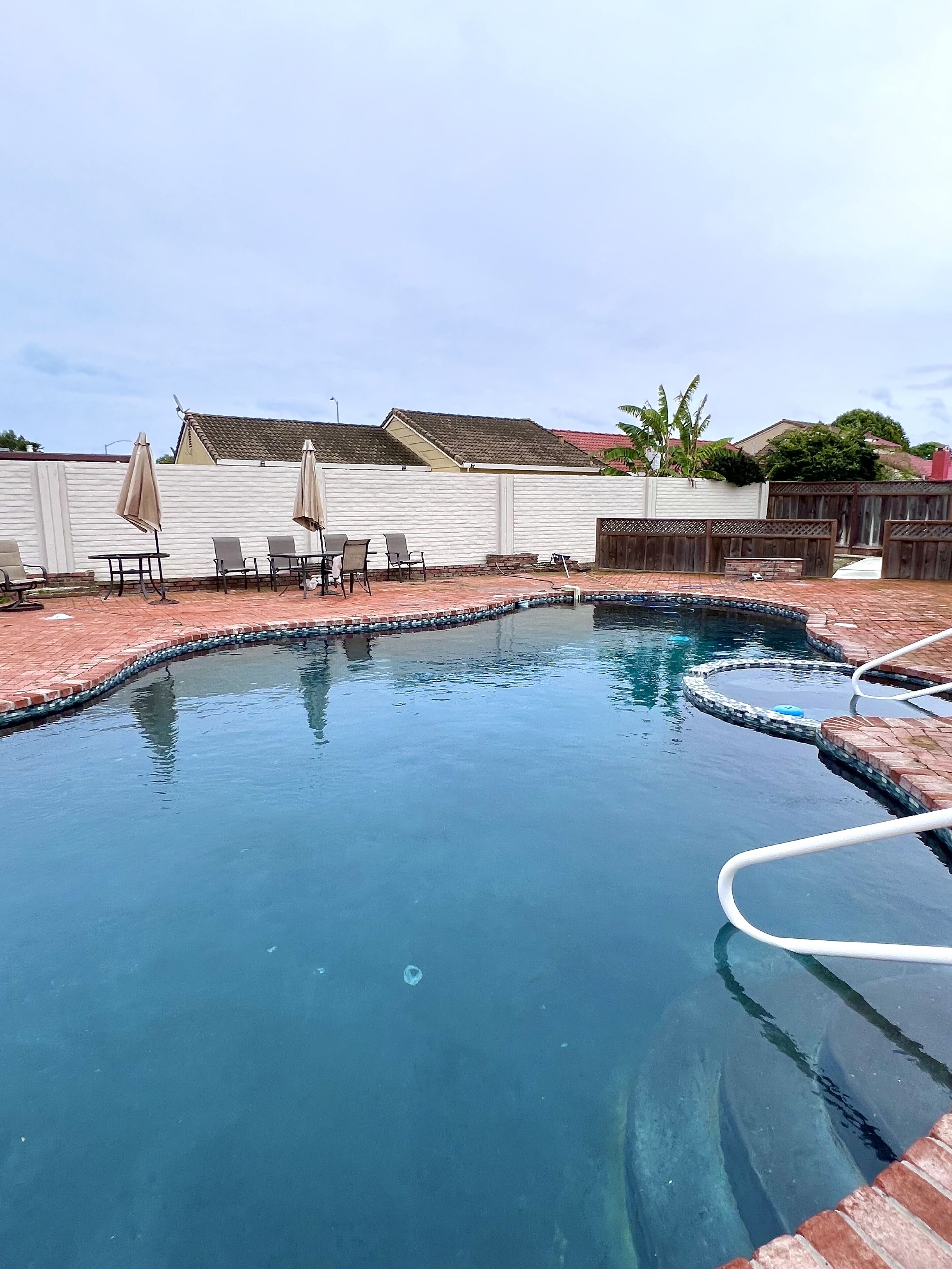 A swimming pool with a brick border, surrounded by a white fence, and includes outdoor seating with umbrellas.