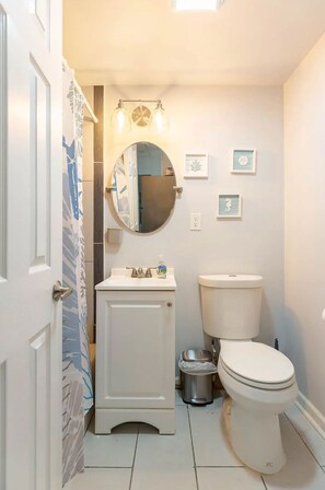Une salle de bain avec des toilettes, un lavabo et un miroir. Les murs sont ornés de tableaux encadrés, et il y a un rideau de douche à motifs de feuilles.