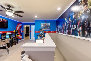 Een kamer met een bureau, gameconsoles en een muurschildering van superhelden.