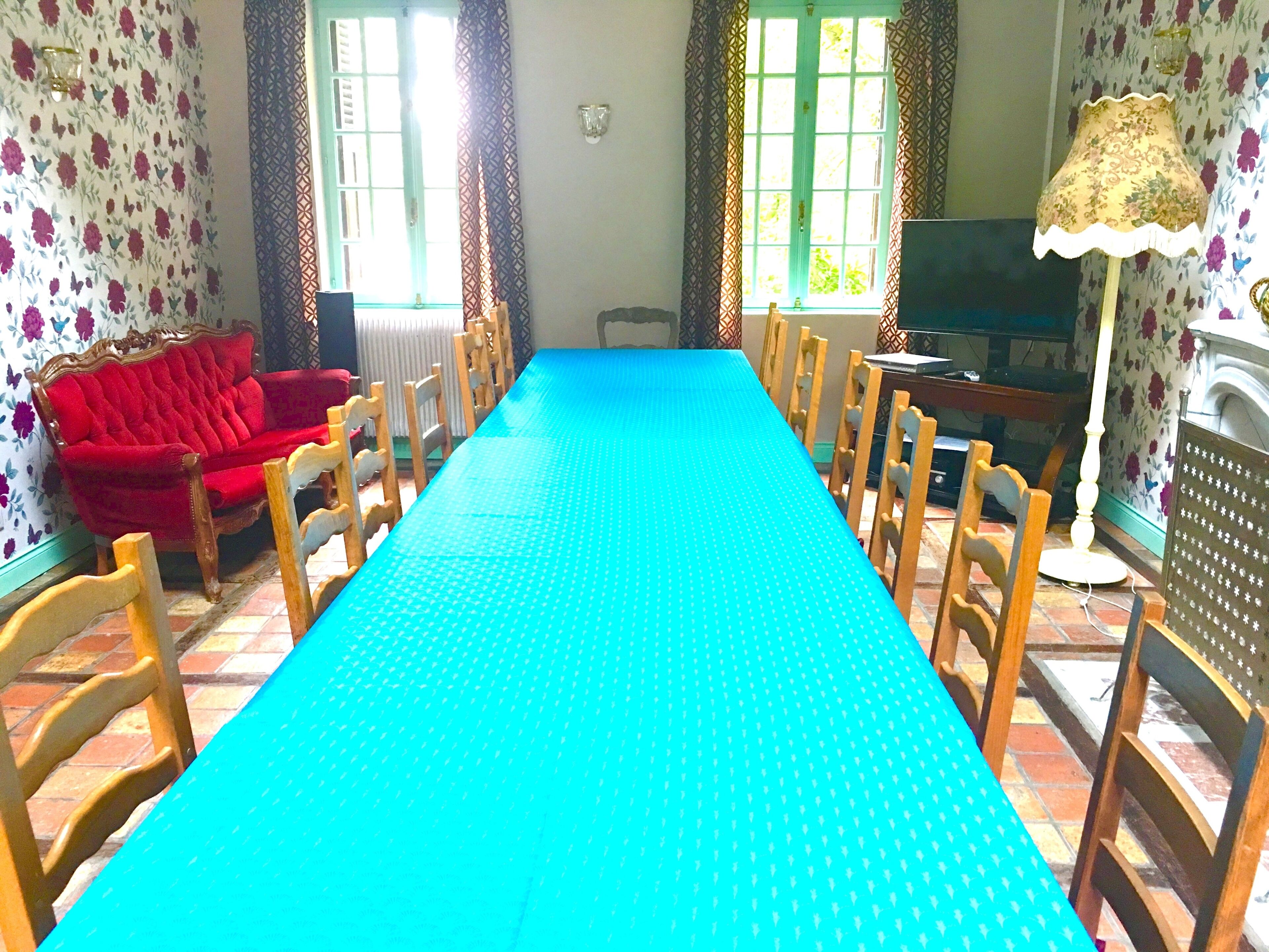 Een eetkamer met een lange tafel, stoelen, een rode bank en een televisie.