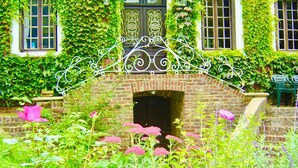 Una chimenea de ladrillo con un arco metálico decorativo encima, rodeada de follaje exuberante y plantas en flor.