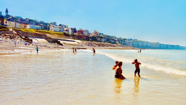 Una playa con gente caminando y jugando en el agua, una costa rocosa y edificios al fondo.