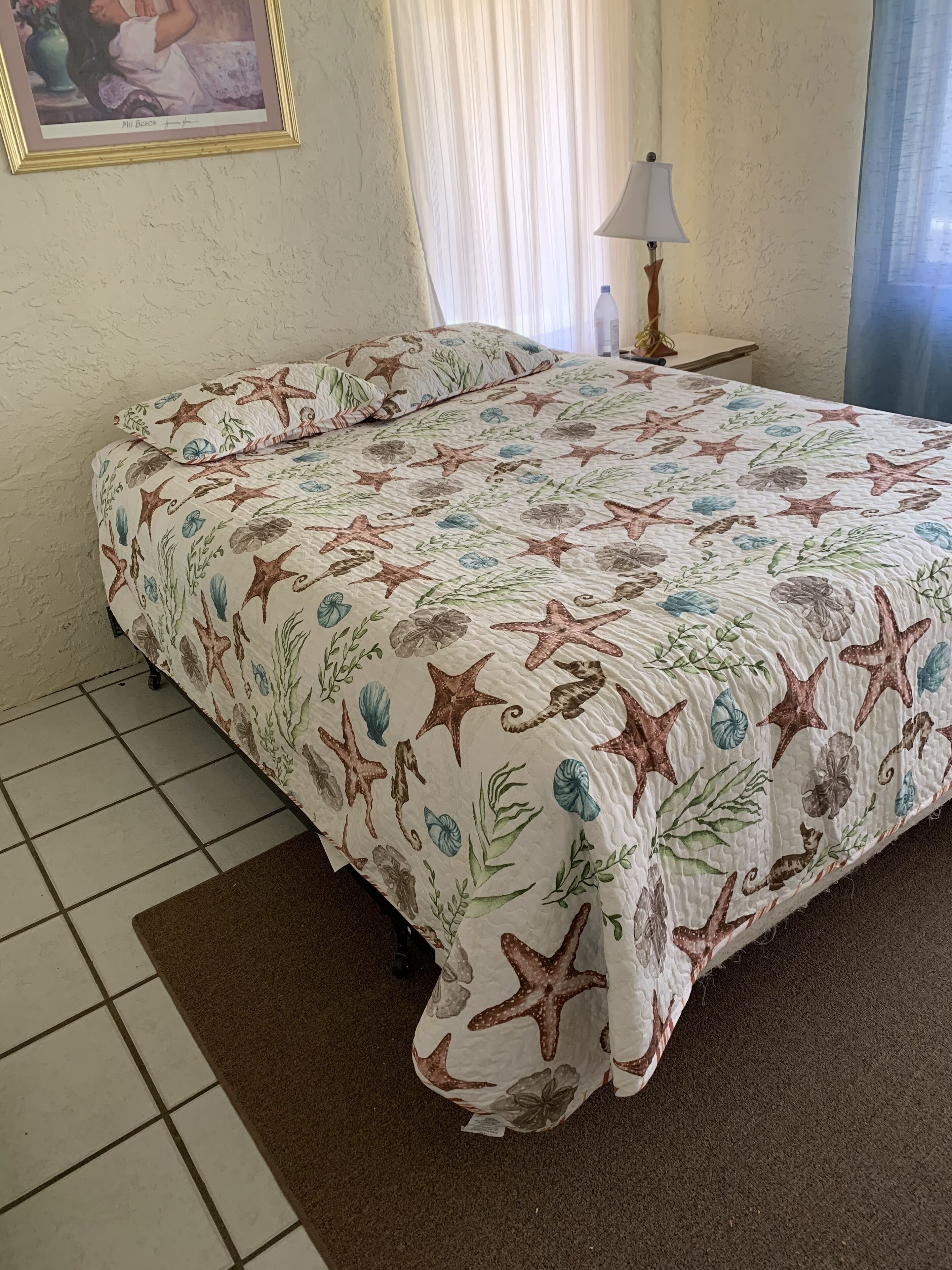 A bedroom with a bed covered in a starfish and seashell patterned bedspread, a small bedside table with a lamp, and a framed picture on the wall.