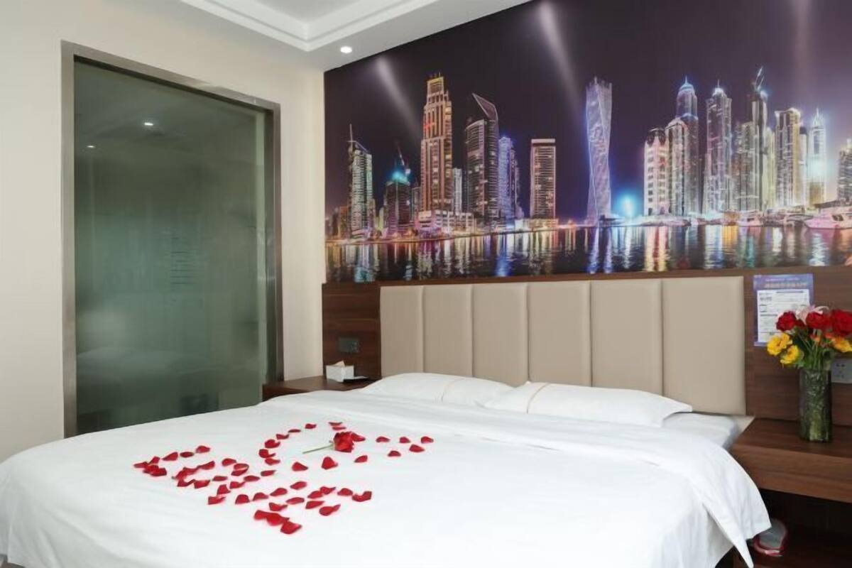 A hotel room with a bed, a night cityscape mural, and a vase with flowers on the nightstand.