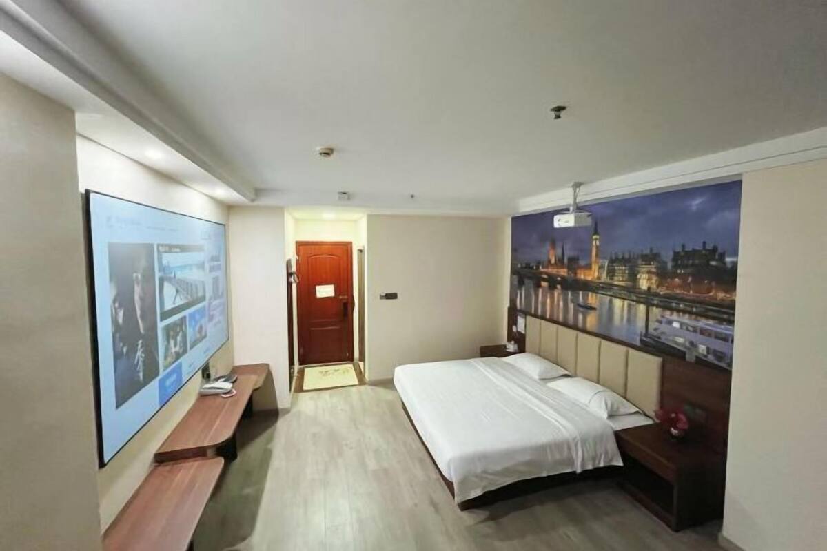 A hotel room with a bed, a TV, a bench, a door, and a cityscape mural on the wall.