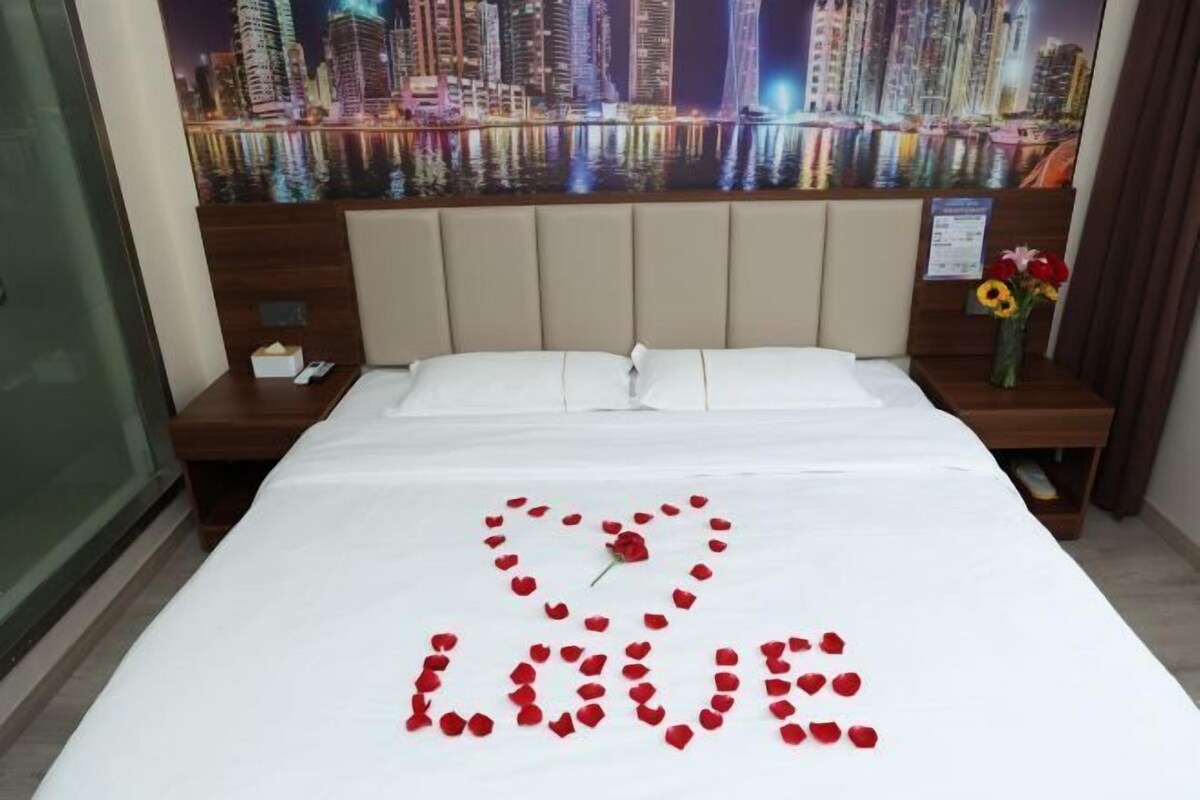 A hotel room with a bed, a nightstand, a vase with flowers, and a cityscape view.