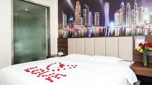 Quarto de hotel com cama, um mural da paisagem urbana, céu noturno e um vaso com flores.