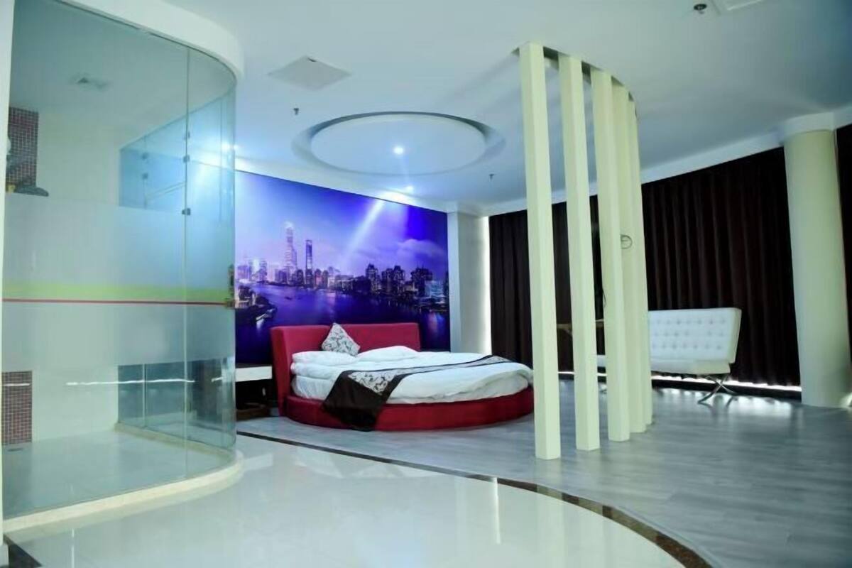 A modern bedroom with a curved glass wall, a cityscape mural, a circular ceiling light, and a red bed.