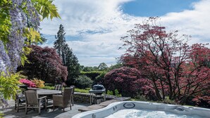 A hot tub surrounded by outdoor furniture, a dining table, and lush greenery.