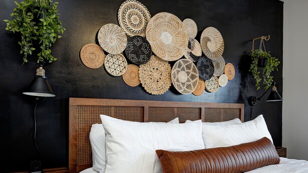 Un dormitorio con cama, almohadas blancas, una almohada decorativa marrón, cabecera, una obra de arte tejida en la pared y dos plantas en macetas.
