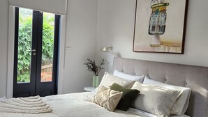 A bedroom with a bed, a door leading to a garden, a framed artwork on the wall, and a nightstand with a vase. - Jayda- **A split level home with a magical ambience ** (Daylesford)