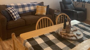 Ein gemütliches Wohnzimmer mit einem flauschigen Fellteppich, einer karierten Tischdecke, einem Ledersessel und einem Esstisch mit einer Holzdekoration.