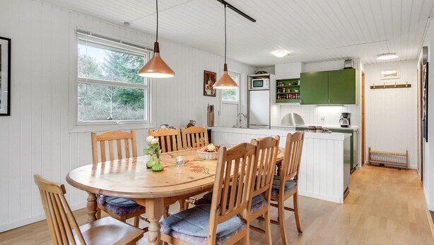 A dining area with a wooden table and chairs, a kitchen with green cabinets, and a view of trees through the window. - Beautiful home in Saltum with kitchen (Saltum)