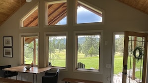 Amplia habitación con techos altos, ventanas grandes que dan a un paisaje cubierto de hierba y una terraza de madera afuera.