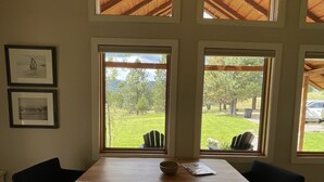 Un comedor con mesa y sillas de madera, ventanas amplias con vistas a un paisaje verde y cuadros en la pared.