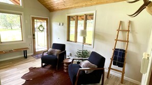 Amplia habitación con vigas de madera en el techo, un ventanal grande, un banco de madera, dos sillones, una mesa de centro, un tapiz de cuero y un cornamenta de ciervo montada.
