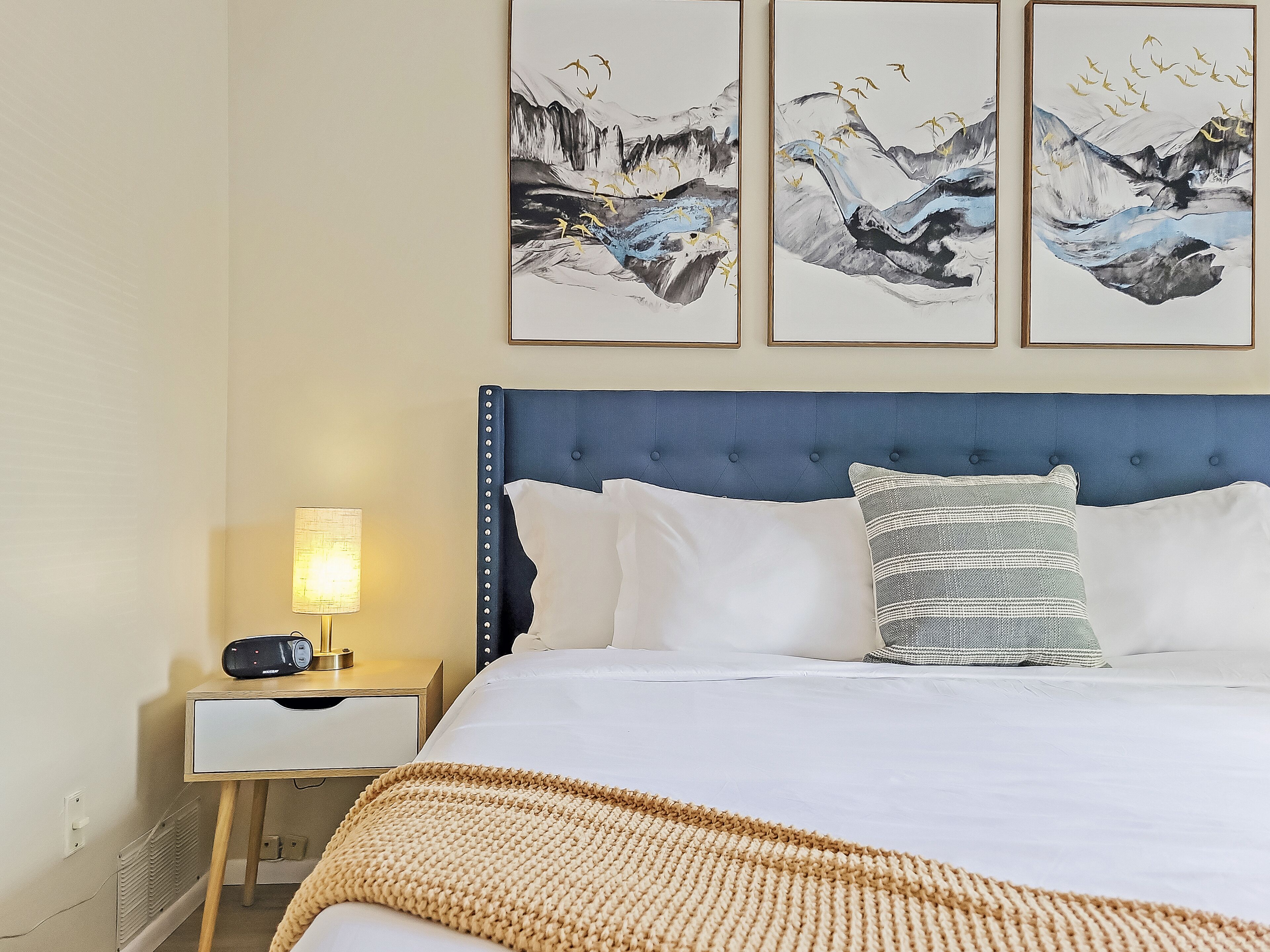 Een netjes opgemaakt bed met een blauw hoofdeinde, witte kussens en een beige gebreide plaid. Er staat een nachtkastje met een lamp en een radio. Drie ingekaderde schilderijen van bergen en vogels hangen aan de muur.