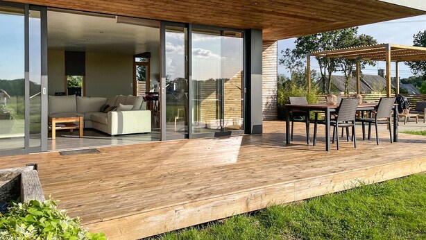 A modern house with a wooden deck, a glass sliding door leading to a living room, and an outdoor dining area with chairs and a table. - 3 bedroom awesome home in Plélan-le-Petit (Plélan-le-Petit)