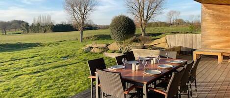 An outdoor dining area with a wooden table and chairs, set against a backdrop of a grassy field and trees.