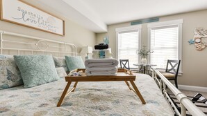 Une chambre à coucher avec un lit, des oreillers, une table de chevet avec des articles de toilette, une citation encadrée au mur et une fenêtre avec des stores.