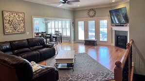 A spacious living room with a black leather couch, a coffee table, and a fireplace. There is a dining area with a table and chairs, and a large window providing natural light.