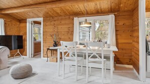 Een gezellig cabin interieur met een houten plafond, een eettafel met witte stoelen, een televisie en een raam met gordijnen.