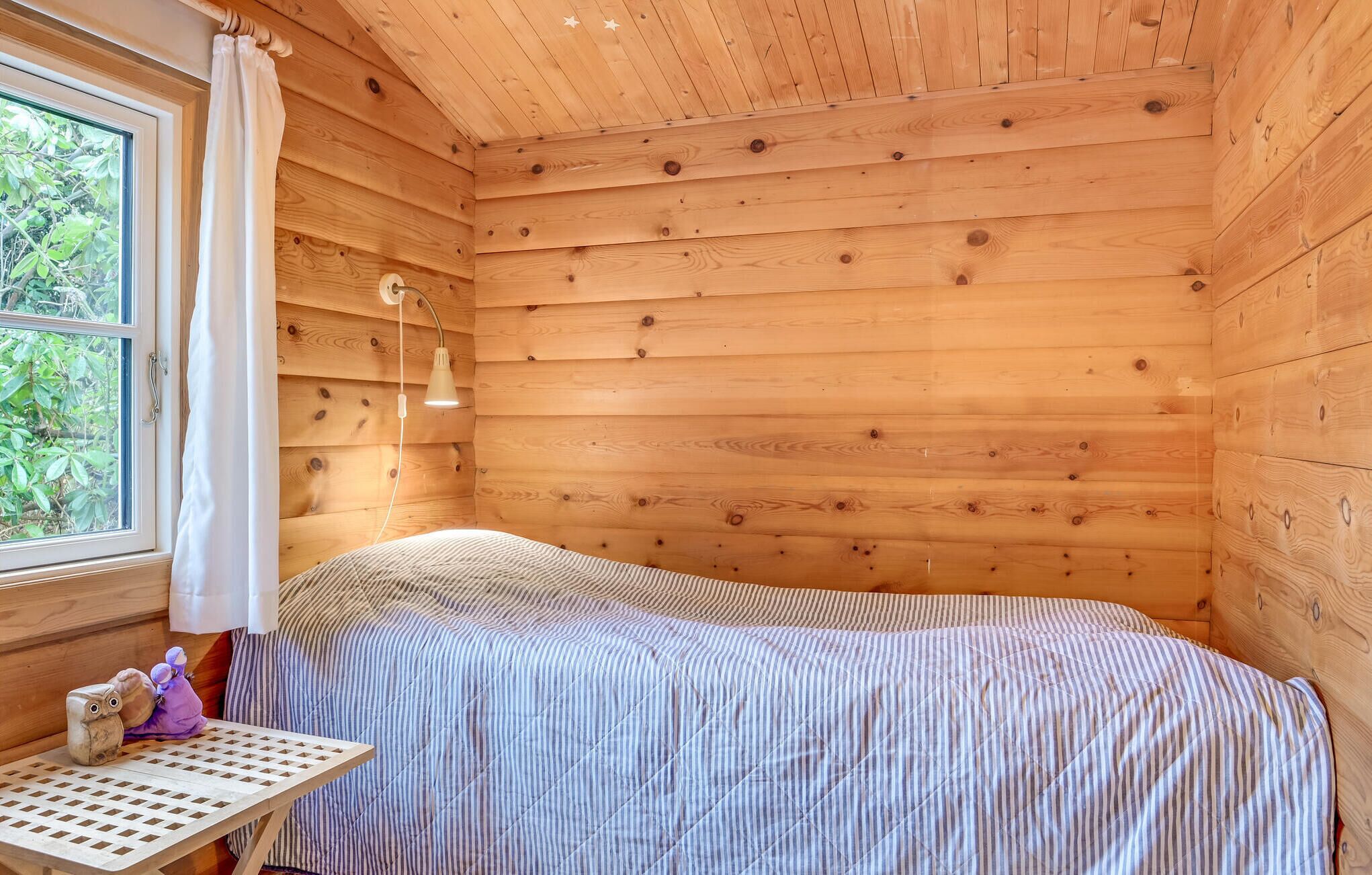 A wooden cabin room with a bed, a table with a lamp, and a window with curtains.