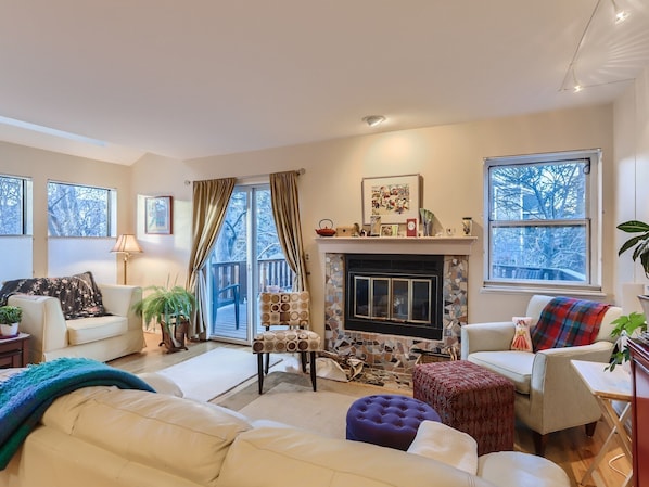 A cozy living room with a fireplace, comfortable seating, and large windows offering a view of the outdoors.